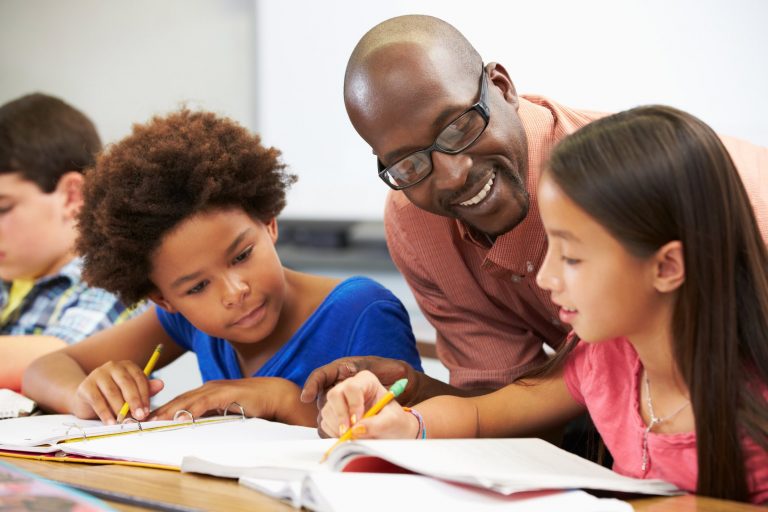 Teacher helping pupils study in the classroom Teacher Helping Pupils Studying At Desks In Classroom Checking Work Smiling